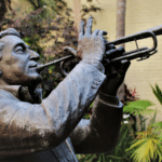 Let’s Connect at NOLA MusiCon 2025 Bronze statue of a man playing the trumpet, surrounded by palm trees and plants, evokes the vibrant spirit of NOLA, with a neutral wall in the background.