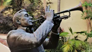 Bronze statue of a man playing the trumpet, surrounded by palm trees and plants, evokes the vibrant spirit of NOLA, with a neutral wall in the background.