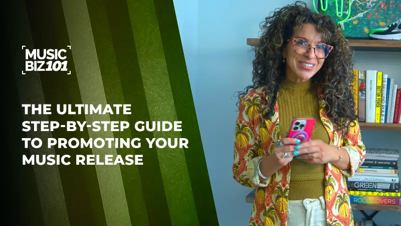 A woman with curly hair and glasses stands holding a phone next to a sign reading "The Ultimate Step-By-Step Guide to Promoting Your Music Release.