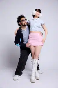A man in glasses and a sweater poses beside a woman wearing earmuffs, a blue gingham crop top, a pink mini skirt, and white lace-up boots, both standing against a plain background.