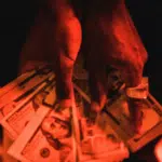 A person wearing a ring holds several spread-out U.S. $100 bills under red lighting, capturing the vibe of success often associated with the music distribution industry.