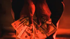 A person wearing a ring holds several spread-out U.S. $100 bills under red lighting, capturing the vibe of success often associated with the music distribution industry.