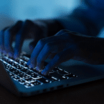 Close-up of hands typing on a laptop keyboard in a dimly lit environment, with the keys illuminated by the laptop’s backlight.