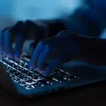 Close-up of hands typing on a laptop keyboard in a dimly lit environment, with the keys illuminated by the laptop’s backlight.