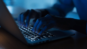 Close-up of hands typing on a laptop keyboard in a dimly lit environment, with the keys illuminated by the laptop’s backlight.