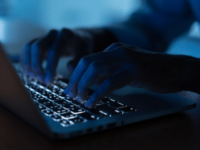Close-up of hands typing on a laptop keyboard in a dimly lit environment, with the keys illuminated by the laptop’s backlight.