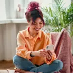 A woman with pink hair sits cross-legged on a chair, smiling as she scrolls through TikTok on her smartphone in a bright, cozy room filled with plants.