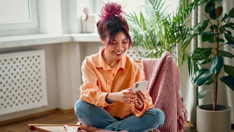 A woman with pink hair sits cross-legged on a chair, smiling as she scrolls through TikTok on her smartphone in a bright, cozy room filled with plants.