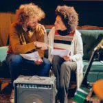 Submit Your Music To Our Dinner Table Jazz Playlist On Spotify Two people sit on a green couch, holding papers and talking about DSPs amid musical instruments, including guitars and a keyboard, in a cozy, dimly lit room.