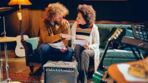 Two people sit on a green couch, holding papers and talking about DSPs amid musical instruments, including guitars and a keyboard, in a cozy, dimly lit room.