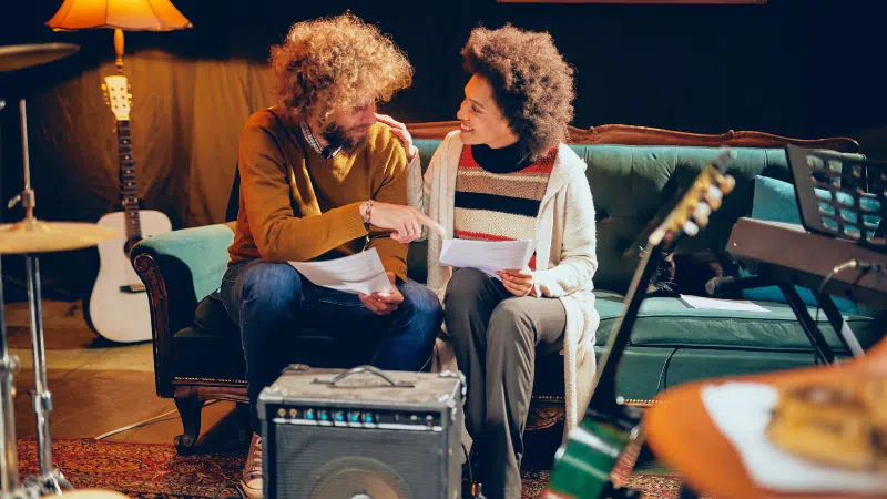 Top Features Artists Love in DSPs and Why They Matter in 2025 Two people sit on a green couch, holding papers and talking about DSPs amid musical instruments, including guitars and a keyboard, in a cozy, dimly lit room.