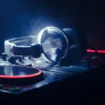 A pair of headphones rests on a DJ mixer with illuminated red and orange lights in a dimly lit environment, setting the scene for some serious playlist pitching.