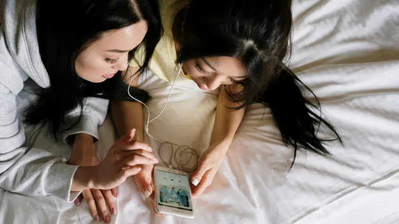 Two people lying on a bed share a pair of earphones, enjoying Apple Music as they look at a smartphone together.