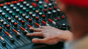 A person adjusts sliders and knobs on a professional audio mixing console, with various colored buttons and controls visible.