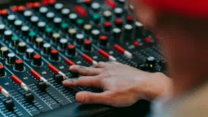 A person adjusts sliders and knobs on a professional audio mixing console, with various colored buttons and controls visible.