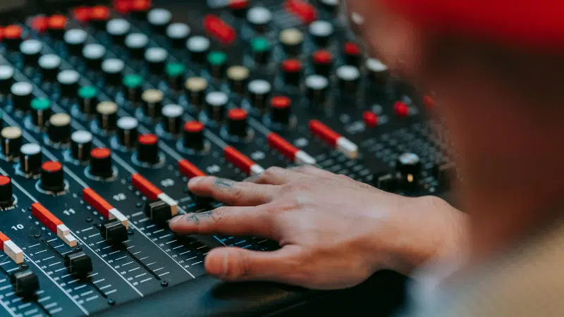 A person adjusts sliders and knobs on a professional audio mixing console, with various colored buttons and controls visible.