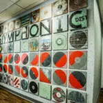 A hallway with wood floors features a symphonic wall art installation made of square tiles with abstract and graphic designs, including the word "SYMPHONIC.