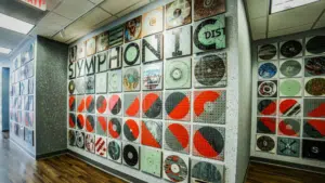 A hallway with wood floors features a symphonic wall art installation made of square tiles with abstract and graphic designs, including the word "SYMPHONIC.