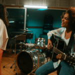 Three musicians perform in a recording studio; one plays keyboard, another sings at a microphone, and a third plays guitar and sings. Drums and audio equipment are visible in the background.