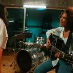 Three musicians perform in a recording studio; one plays keyboard, another sings at a microphone, and a third plays guitar and sings. Drums and audio equipment are visible in the background.