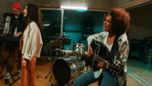 Three musicians perform in a recording studio; one plays keyboard, another sings at a microphone, and a third plays guitar and sings. Drums and audio equipment are visible in the background.