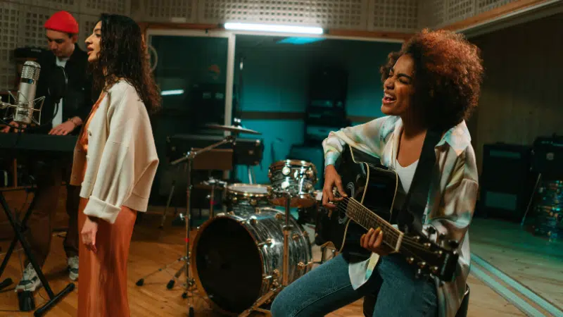 Three musicians perform in a recording studio; one plays keyboard, another sings at a microphone, and a third plays guitar and sings. Drums and audio equipment are visible in the background.