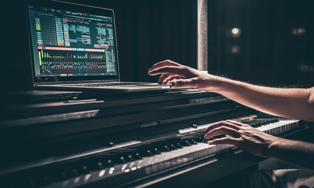Person playing an electronic keyboard with a laptop on top, displaying Meta music production software on the screen in a dimly lit setting.