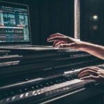 Person playing an electronic keyboard with a laptop on top, displaying Meta music production software on the screen in a dimly lit setting.
