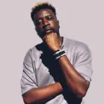 A man stands against a plain background, wearing a light gray T-shirt and bracelets, his hand resting thoughtfully on his chin—capturing the cool, introspective vibe of Afro House style.