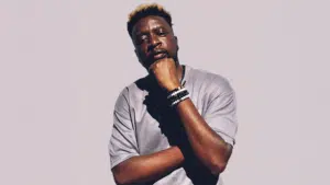 A man stands against a plain background, wearing a light gray T-shirt and bracelets, his hand resting thoughtfully on his chin—capturing the cool, introspective vibe of Afro House style.