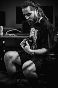 A man with long hair and a beard sits and plays an electric guitar in a recording studio, wearing a t-shirt and shorts. Black and white photo.