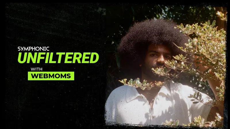A person with a large afro stands partially behind leafy branches; text reads "Symphonic Unfiltered with Webmoms." The scene highlights an unfiltered vibe, blending natural elements with authentic energy.