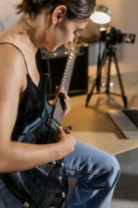 A person in a black sleeveless top and jeans plays an electric guitar while seated at a desk with a laptop, camera, and amplifier in the background, creating content for YouTube for Artists.
