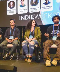 Four panelists sit on stage during an Indie Week music conference, with name tags and a presentation screen displaying their names and roles behind them.