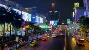 A busy Bangkok city street at night with bright lights, illuminated billboards, tall buildings, cars, buses, and people along the sidewalks.