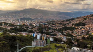 A panoramic view of Medellin with densely packed buildings nestled between green hills and mountains under a cloudy sky at sunset.