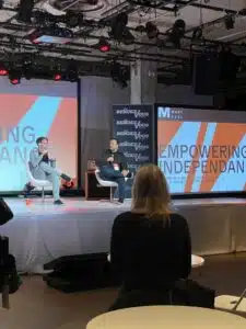 Two speakers sit on stage at a music conference, engaging in discussion with an audience. Bright screens display the words "EMPOWERING INDEPENDENCE" and "RENDEZVOUS.