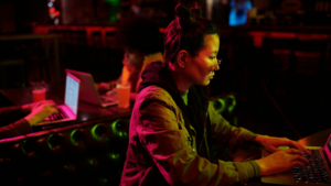 A person wearing glasses works on a laptop in a dimly lit setting, with another person using a laptop in the background—both immersed in their tasks, reflecting the focused work culture at Meta.
