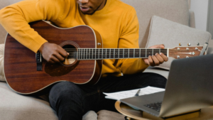 A person in a yellow sweater plays an acoustic guitar while sitting on a couch, with sheet music and an open laptop in front of them, perhaps recording content for YouTube.