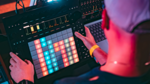 Person using a digital music production controller with colorful LED pads, next to a laptop, in a studio setting.