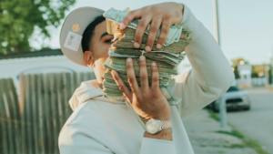 A person in a white hoodie and cap holds multiple stacks of cash, partially obscuring their face, while wearing a watch outdoors—possibly celebrating a big win in music publishing.