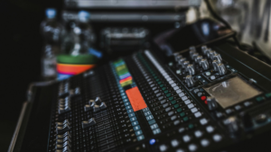 A close-up view of an audio mixing console enhanced by AI, featuring multiple sliders, knobs, colored buttons, and a small display screen. Bottled water and other equipment are visible in the background.