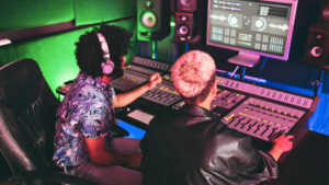 Two people work at a mixing console in a recording studio, adjusting audio levels while viewing sound analytics on a large computer screen.