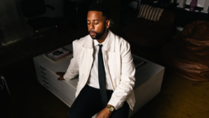 A man in a white jacket and black tie sits with his eyes closed on a white table in a dimly lit room.
