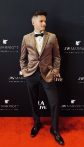 A man in a tan velvet blazer, white dress shirt, black bow tie, and black pants stands on a red carpet in front of a JW Marriott L.A. Live backdrop.