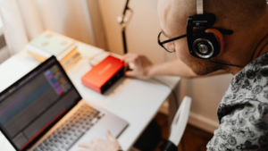 A person wearing headphones uses a laptop and audio interface at a desk, with books, a lamp, and documents from distributors in the background.