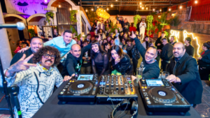 A group of DJs poses for a selfie behind turntables at a lively FIMPro indoor party, with a crowd of people smiling and cheering in the background under string lights.