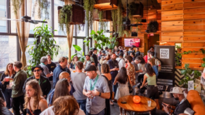 A large group of people socialize at an indoor SXSW event space with wooden walls, hanging plants, and natural light streaming through large windows.