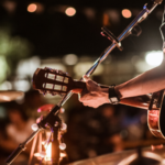A musician plays an acoustic guitar on stage under string lights, with a microphone nearby and a blurred audience in the background—perfect for sharing a live performance on YouTube.