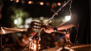 A musician plays an acoustic guitar on stage under string lights, with a microphone nearby and a blurred audience in the background—perfect for sharing a live performance on YouTube.